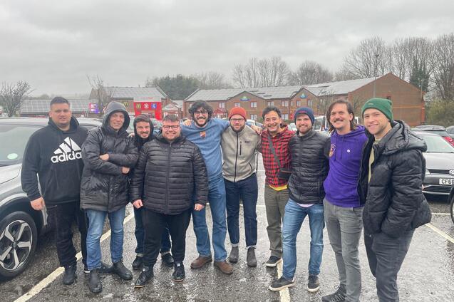 Gruppe von zehn M&auml;nnern in wetterfester Kleidung l&auml;chelt gemeinsam auf einem Parkplatz bei bew&ouml;lktem Himmel.