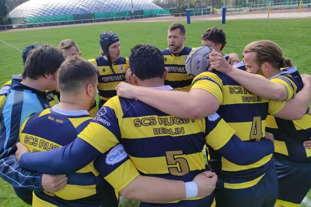 Eine Gruppe von Rugbyspielern in gestreiften Trikots bildet einen Kreis auf einem gr&uuml;nen Spielfeld.