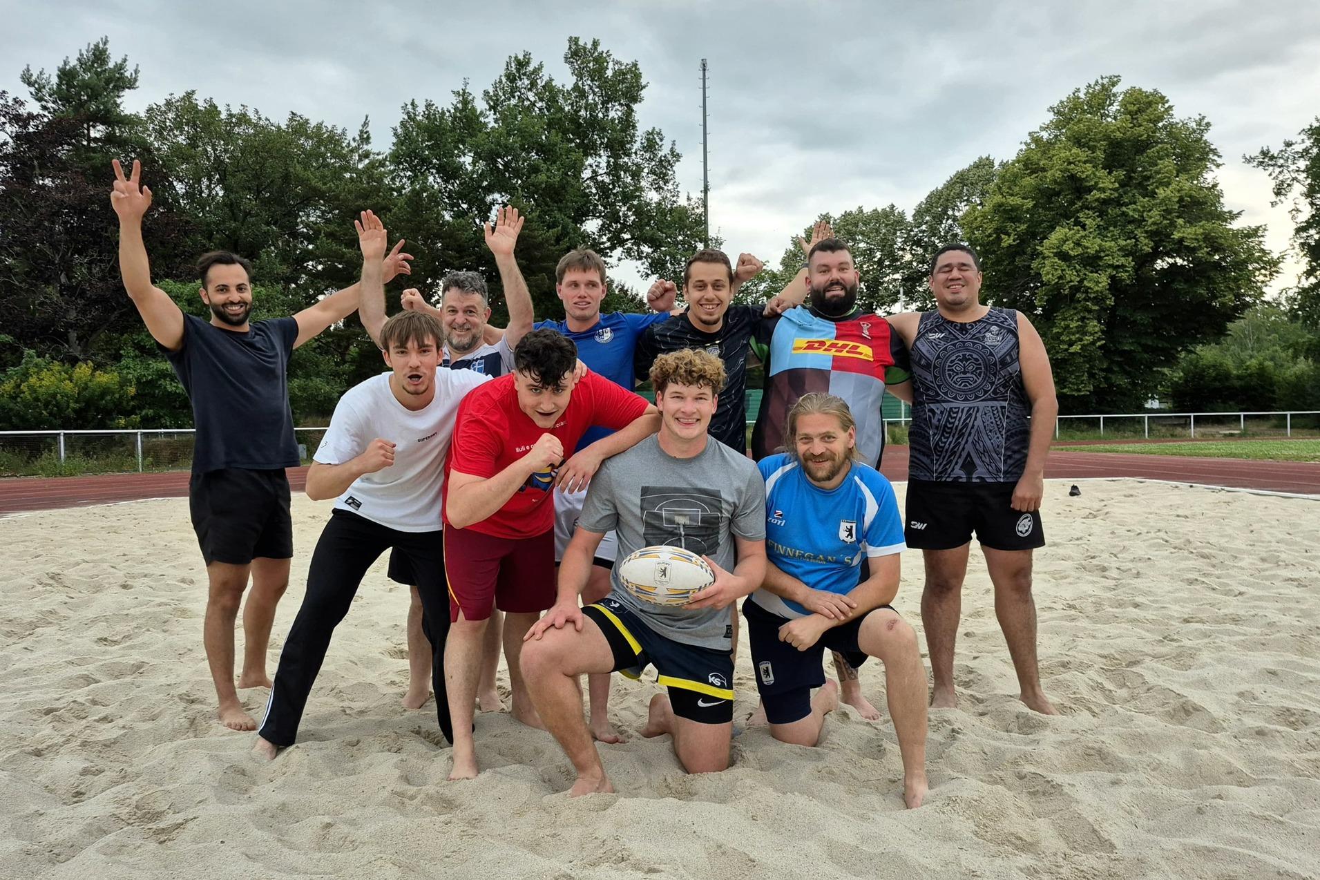 Gruppenshot von zehn Rugbyspielern auf einem Sandfeld, einige posieren, w&auml;hrend andere jubeln.