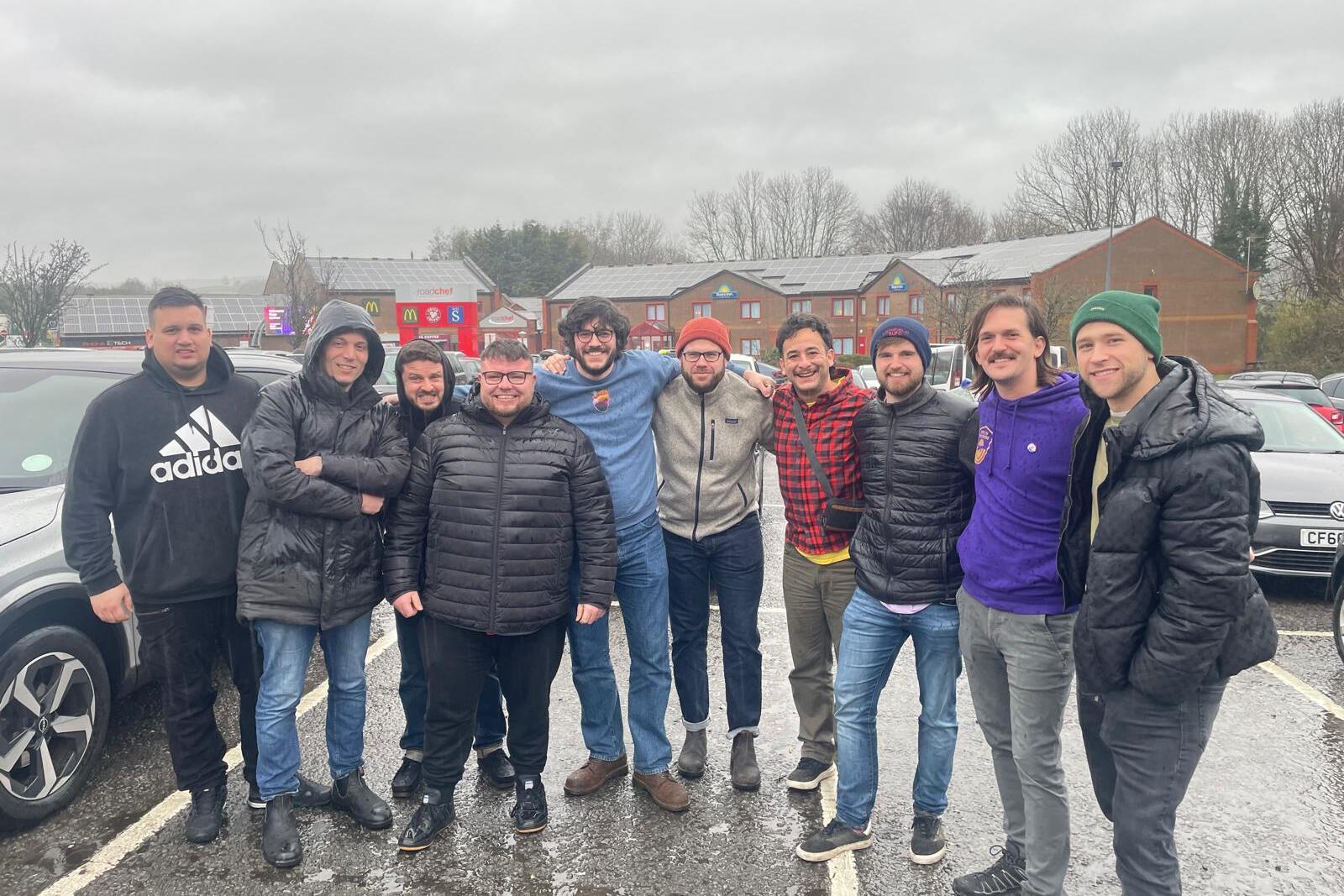 Gruppe von zehn M&auml;nnern in wetterfester Kleidung l&auml;chelt gemeinsam auf einem Parkplatz bei bew&ouml;lktem Himmel.