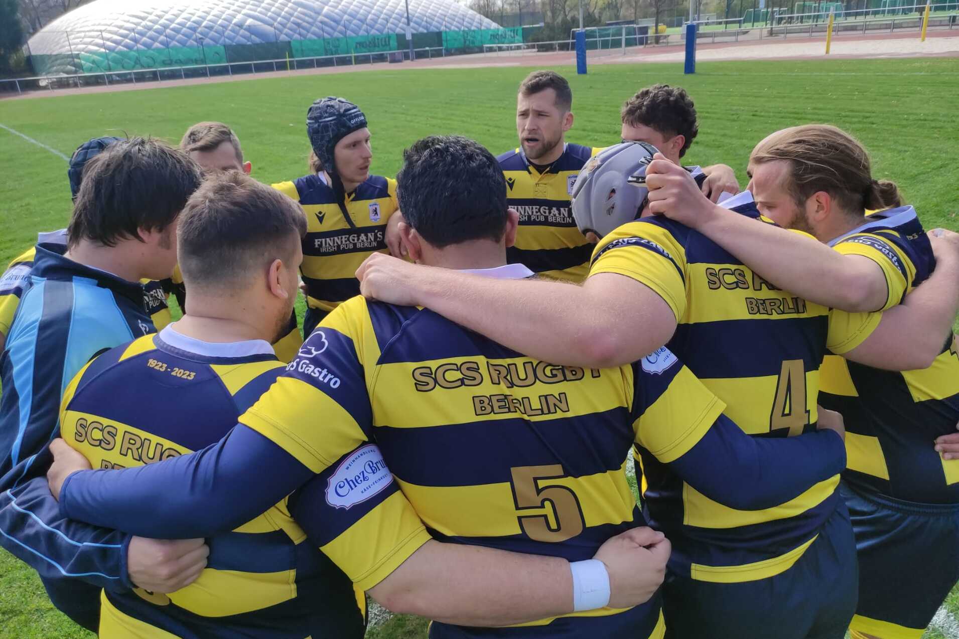 Eine Gruppe von Rugbyspielern in gestreiften Trikots bildet einen Kreis auf einem gr&uuml;nen Spielfeld.