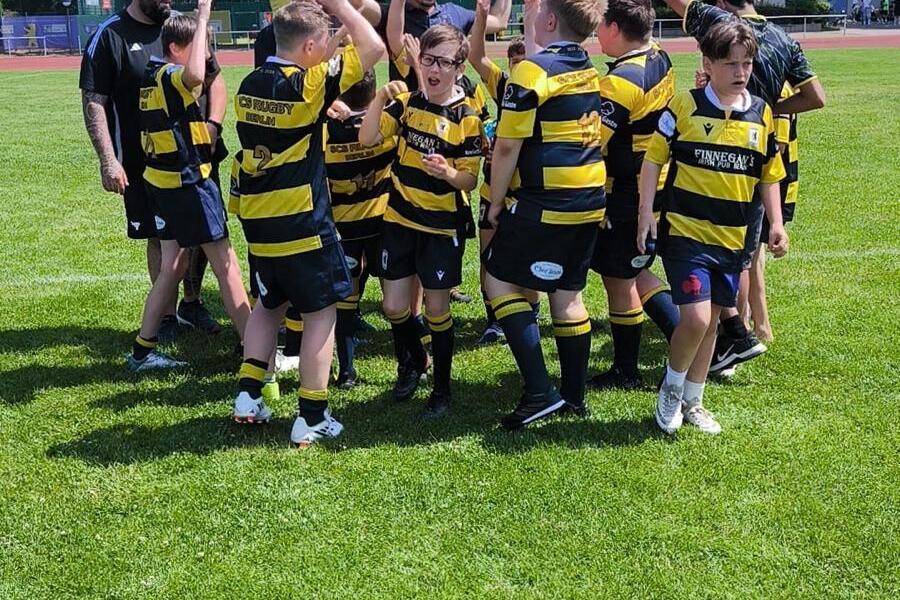 Gruppe von Jugendlichen in gestreiften Trikots feuert sich auf dem Rasenplatz an, während ein Trainer dabei zusieht.