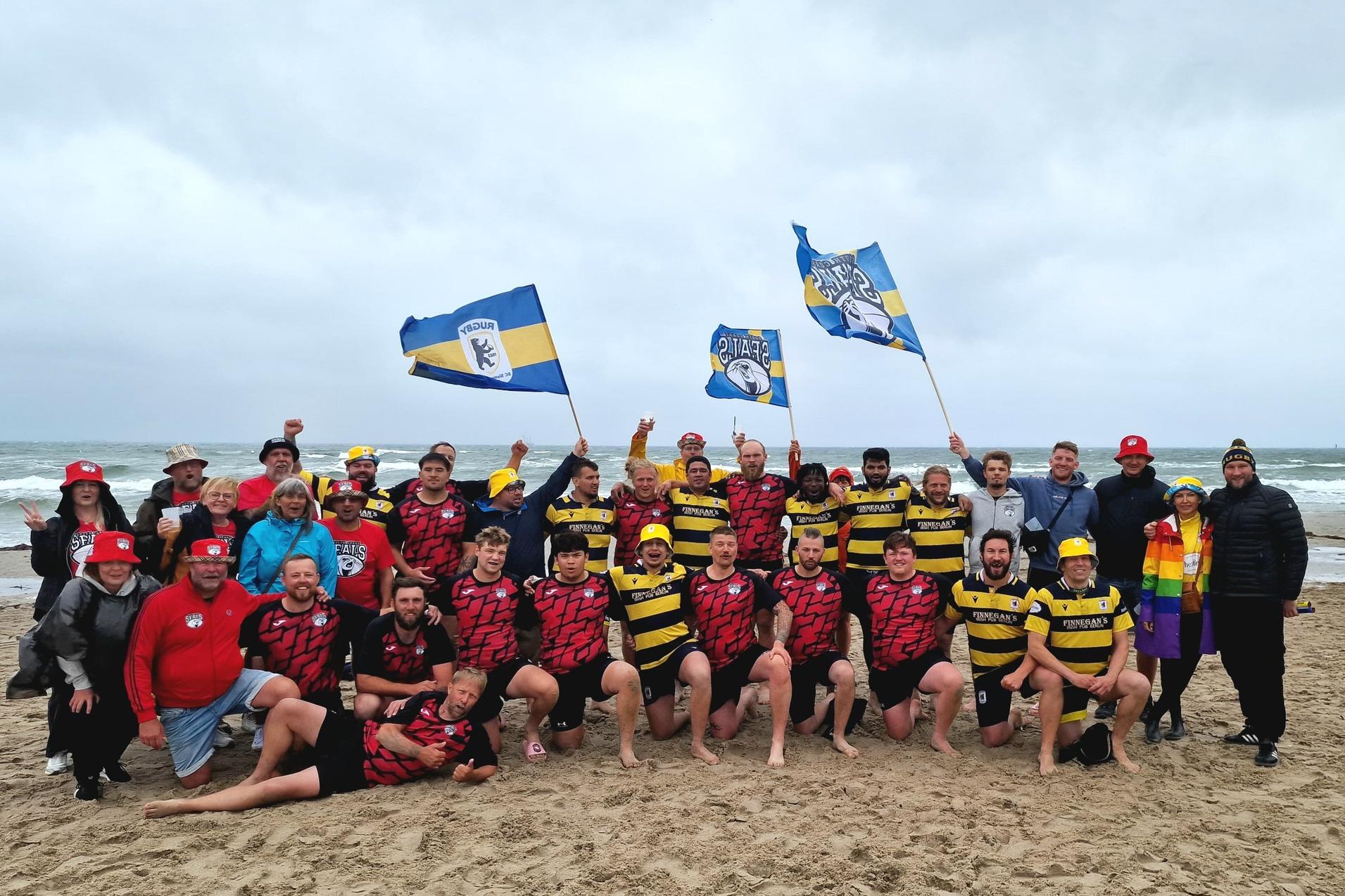 Gruppenshot von Rugbyspielern und Fans am Strand, die Flaggen schwenken und gemeinsam feiern.