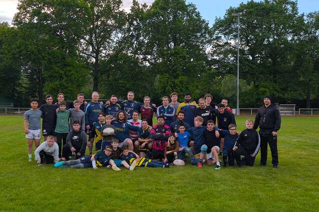 Gruppenfoto von Spielern und Trainern auf einem Rasenplatz nach dem Rugby-Training, umgeben von Bäumen.