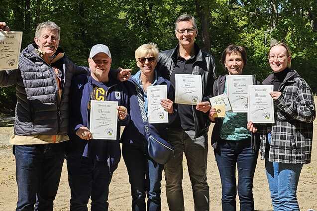 Sechs Personen stehen im Freien und halten Urkunden in den Händen, umgeben von grünen Bäumen und sandigem Boden.