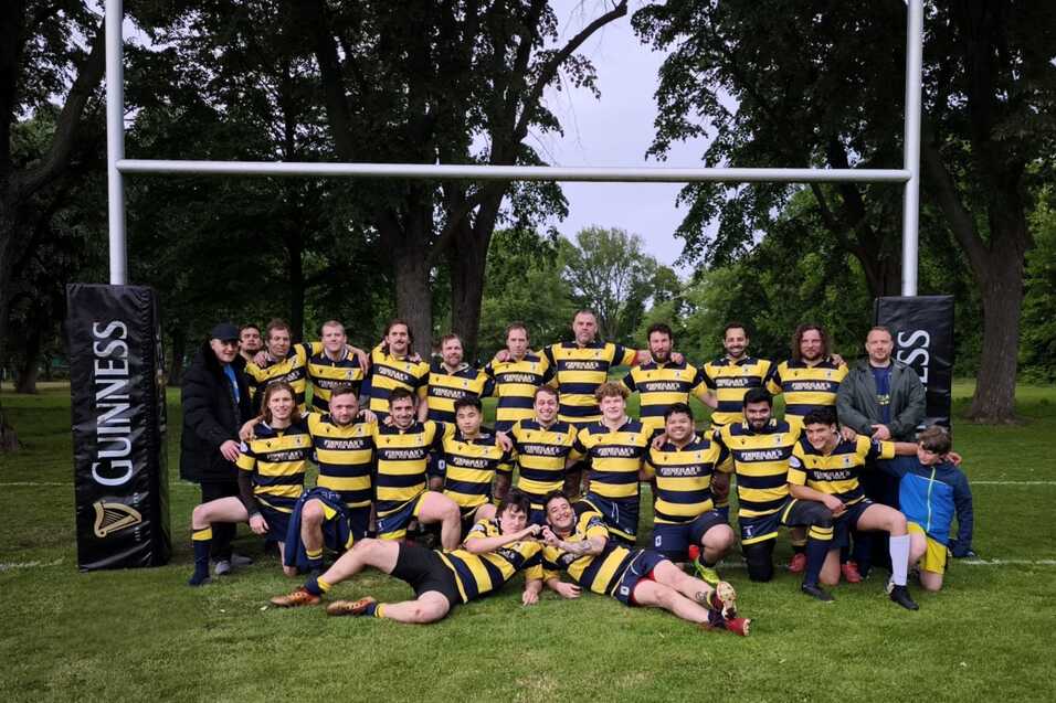 Gruppenshot einer Rugbymannschaft in gestreiften Trikots vor den Torpfosten in einem Park. Emma von hinten links steht und l&auml;chelt.