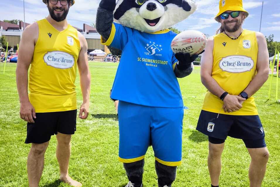 Drei Personen stehen auf einer gr&uuml;nen Wiese, zwei im Sportoutfit und ein Maskottchen mit Rugbyball in der Mitte.