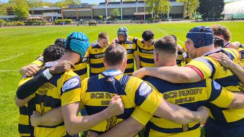 Rugby-Team in gelb-blauen Trikots bildet eine huddle auf einem grünen Platz unter blauem Himmel.