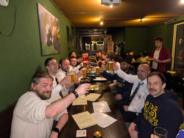 Gruppierung von Männern am Tisch in einer Bar, die mit Biergläsern anstoßen. Hintergrund mit grüner Wand und Bildern.