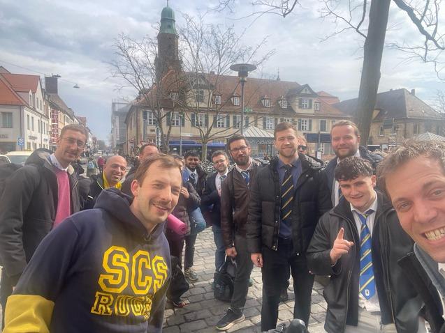 Gruppe von Männern posiert lächelnd auf einer Straße, historische Gebäude und Bäume im Hintergrund, bewölkter Himmel.