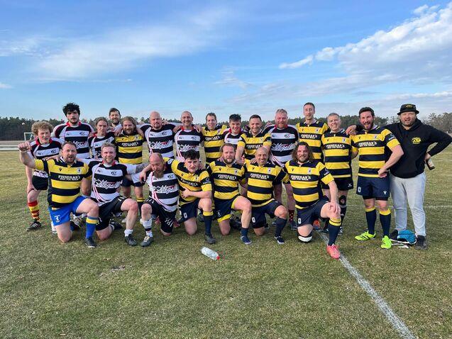 Gruppenfoto von zwei Rugbyteams in einheitlicher Kleidung auf einem Sportfeld, umgeben von Natur und blauem Himmel.