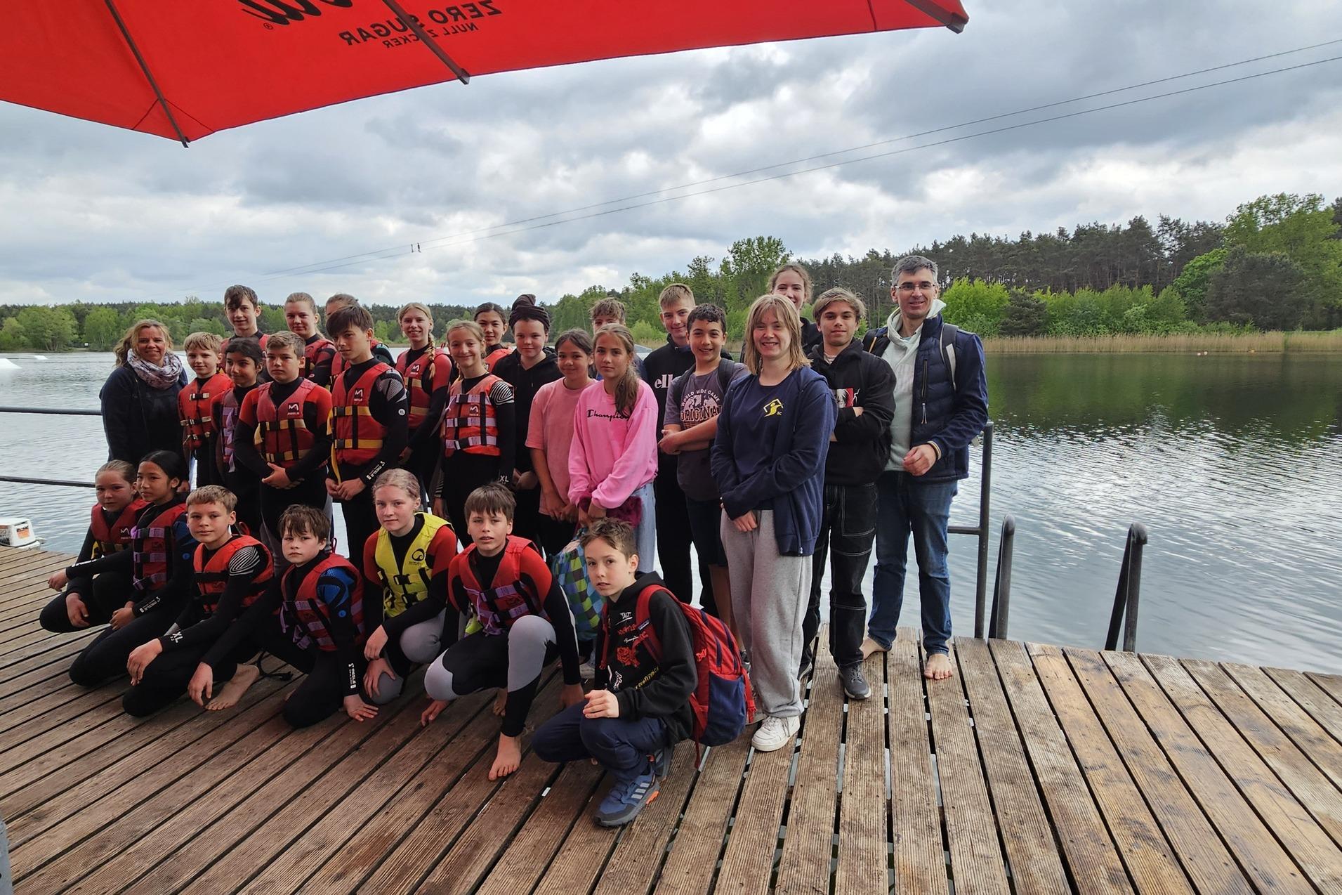 Gruppe von Jugendlichen in Schwimmwesten steht am Ufer eines Sees, umgeben von Bäumen und unter einem roten Sonnenschirm.