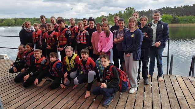 Gruppe von Jugendlichen in Schwimmwesten steht am Ufer eines Sees, umgeben von Bäumen und unter einem roten Sonnenschirm.