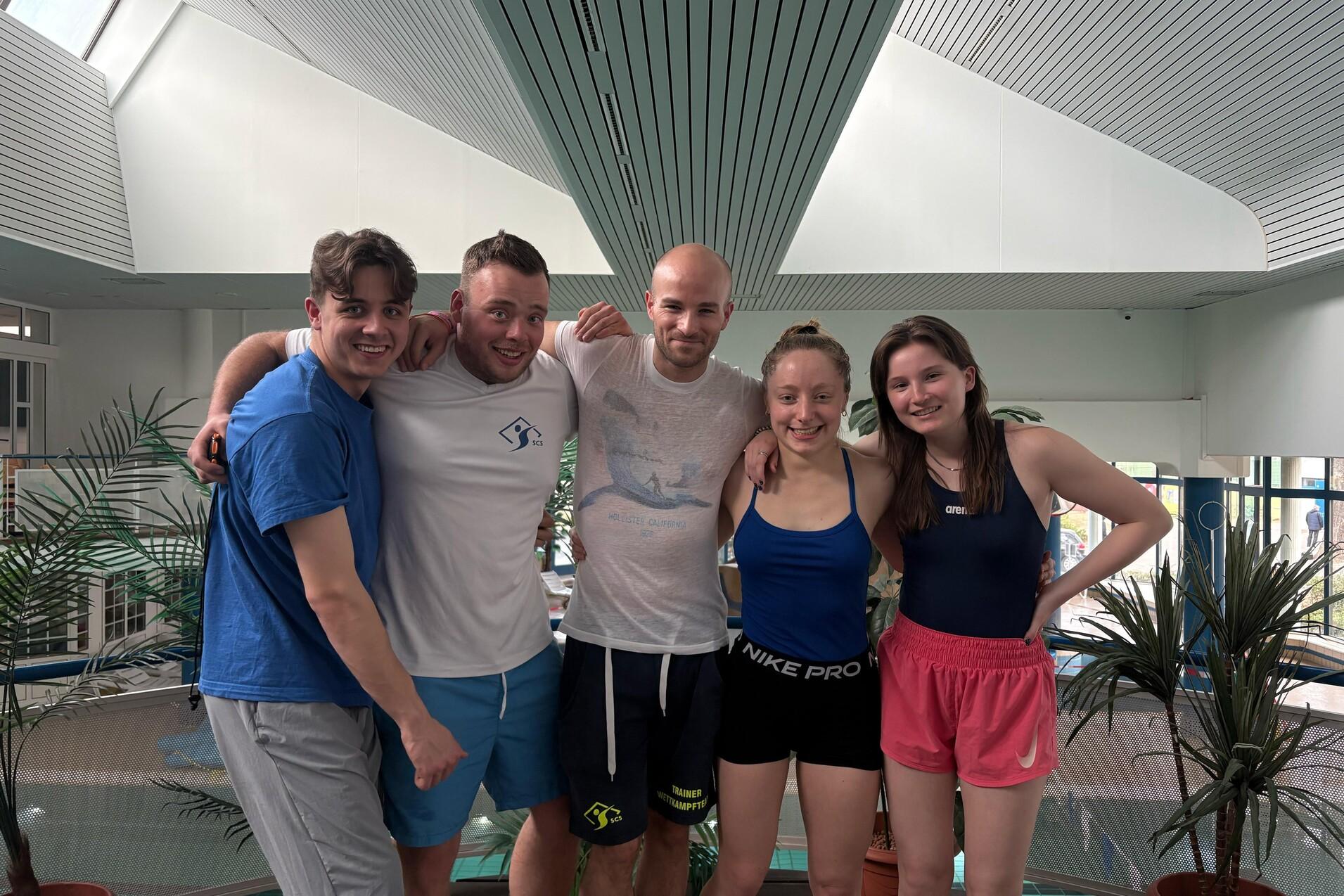 Fünf junge Erwachsene stehen lächelnd mit Arm in Arm in einem Schwimmbadbereich, umgeben von Pflanzen.