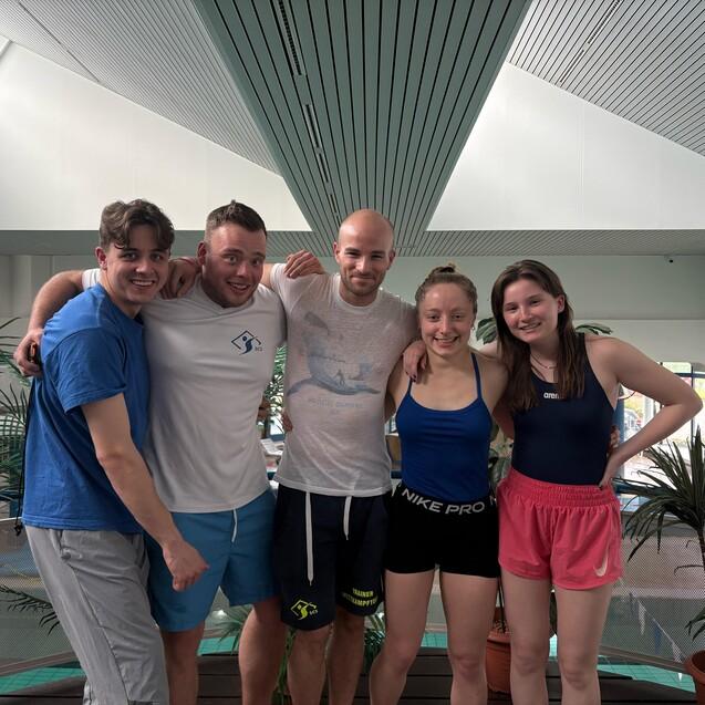 F&uuml;nf junge Erwachsene stehen l&auml;chelnd mit Arm in Arm in einem Schwimmbadbereich, umgeben von Pflanzen.