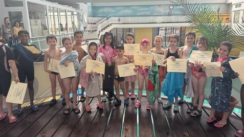 Gruppenszene von Kindern in Badebekleidung mit Urkunden an einem Schwimmbad, umgeben von einer farbigen Wand.