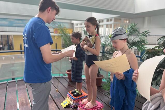 Kinder in Schwimmbekleidung stehen auf einem Podest und halten Papiere, während ein Trainer ihnen Anweisungen gibt.