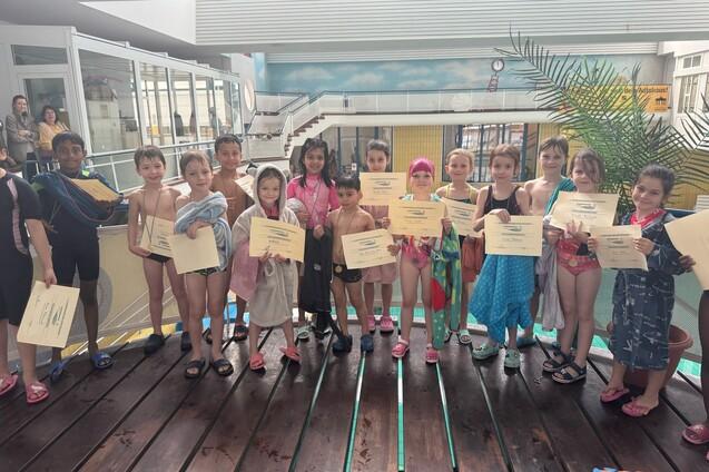 Gruppenszene von Kindern in Badebekleidung mit Urkunden an einem Schwimmbad, umgeben von einer farbigen Wand.