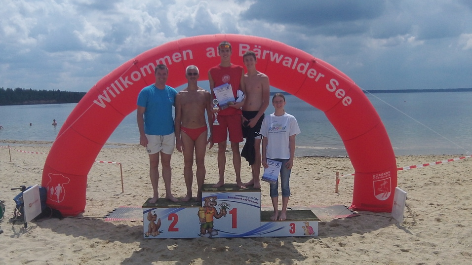 F&uuml;nf Personen stehen auf dem Siegerpodest am B&auml;rwalder See, umringt von Sandstrand und Wasser.