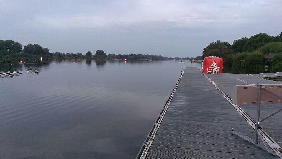 Blick auf einen ruhigen See mit einem Holzsteg und einer roten Aufbewahrungshütte am Ufer.