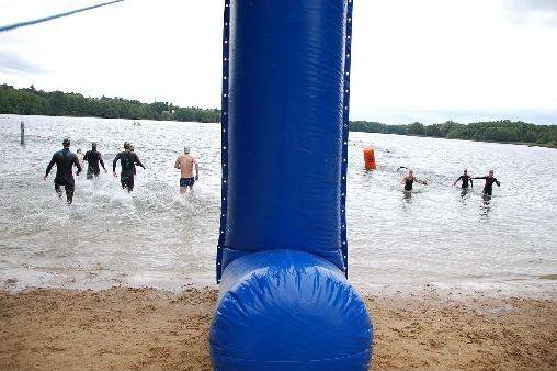 Gruppenschwimmer in Neoprenanzügen laufen ins Wasser, während ein blauer Aufsteller am Ufer steht.