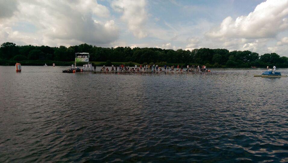Gruppe von Menschen steht auf einem Floß im Wasser, umgeben von einer ruhigen Landschaft mit Bäumen und Wolken.