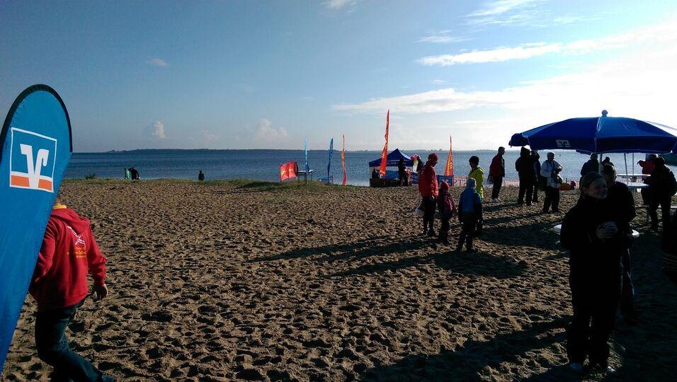 Strand mit mehreren Personen, bunten Fahnen und einem Sonnenschirm, im Hintergrund ein Gew&auml;sser unter blauem Himmel.