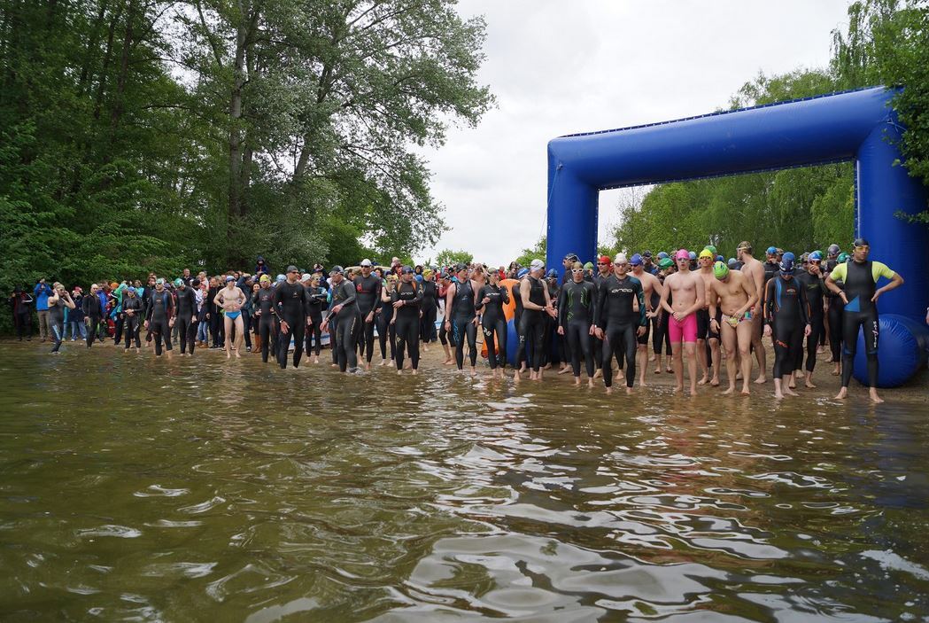 Gruppenschwimmer in Neoprenanz&uuml;gen stehen im flachen Wasser, bereit f&uuml;r den Wettkampf unter einem Startbogen.