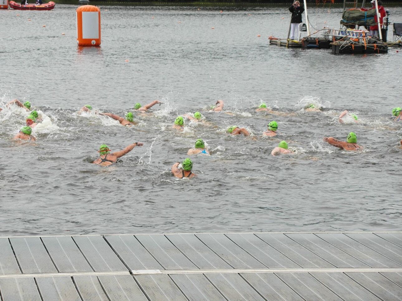 Gruppen von Schwimmern mit gr&uuml;nen Badekappen beim Wettkampf in einem See, im Hintergrund ein Bootssteg.