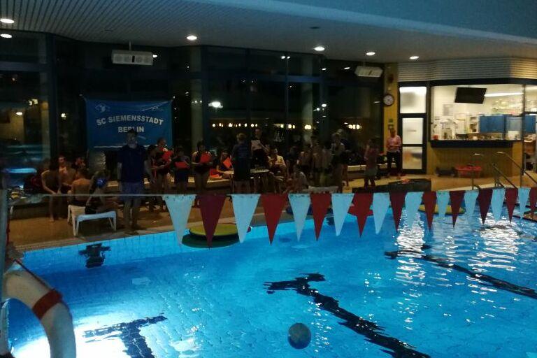Blick auf ein beleuchtetes Schwimmbad mit Schwimmenden und Menschen am Rand, im Hintergrund ein Banner des SC Siemensstadt.