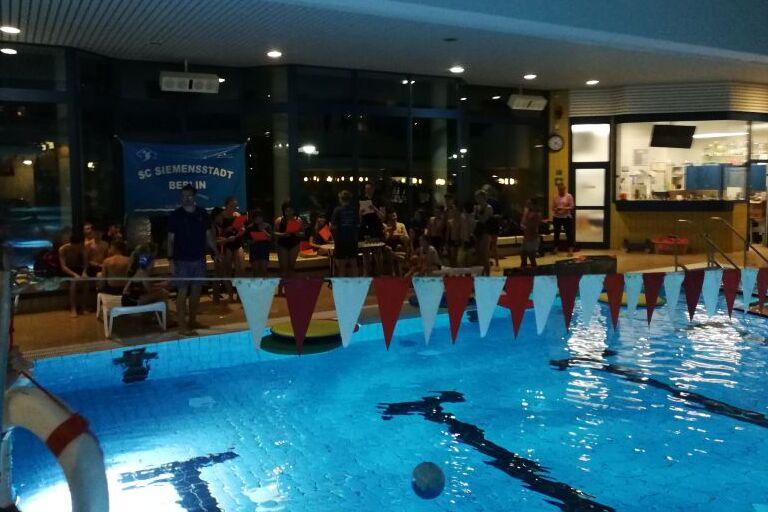 Blick auf ein beleuchtetes Schwimmbad mit Schwimmenden und Menschen am Rand, im Hintergrund ein Banner des SC Siemensstadt.