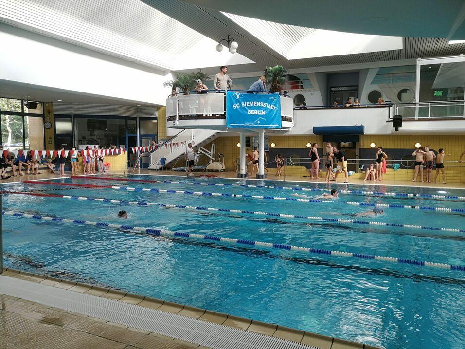 Innenansicht eines Schwimmbades mit Schwimmern und Zuschauern an der Beckenrand. Im Hintergrund eine Trib&uuml;ne mit Banner.