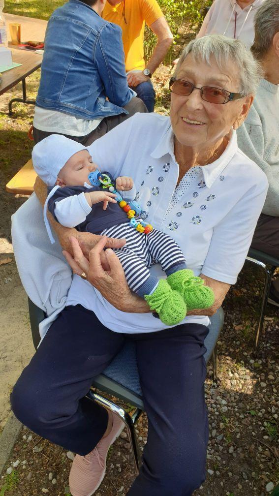 Eine ältere Frau hält ein schlafendes Baby in den Armen, umgeben von Personen in einer sonnigen Umgebung.