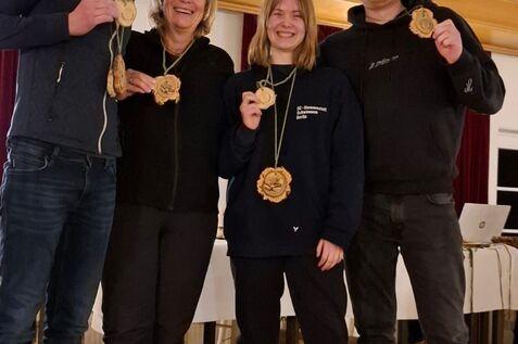 Vier Personen stehen zusammen und halten Medaillen in der Hand, vor einer Holzverkleidung und Tischen.