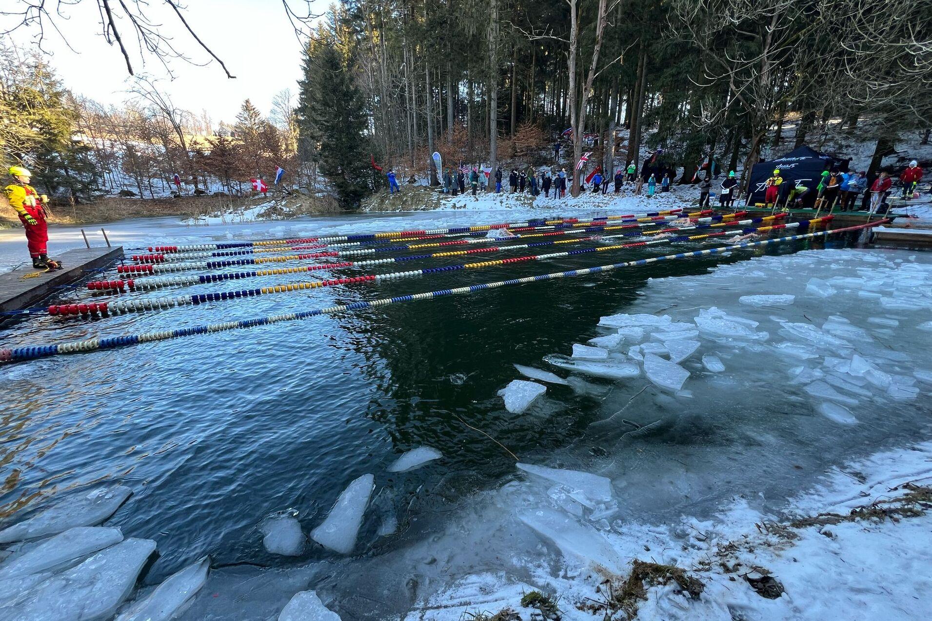 Personen beobachten einen Schwimmwettbewerb in einem kalten Gewässer, umgeben von Eis und schneebedeckten Bäumen.