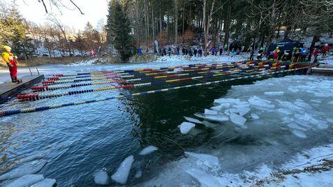 Personen beobachten einen Schwimmwettbewerb in einem kalten Gewässer, umgeben von Eis und schneebedeckten Bäumen.