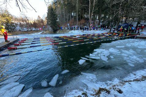 Personen beobachten einen Schwimmwettbewerb in einem kalten Gewässer, umgeben von Eis und schneebedeckten Bäumen.