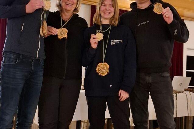 Vier Personen stehen zusammen und halten Medaillen in der Hand, vor einer Holzverkleidung und Tischen.