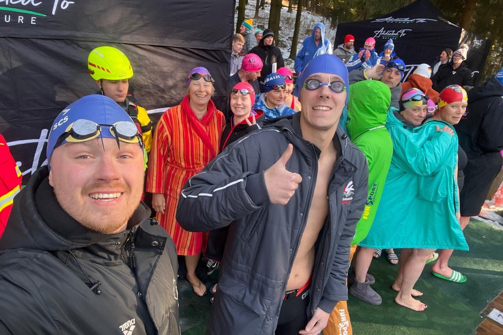 Gruppe von Schwimmern in Badebekleidung und farbenfrohen &Uuml;berbekleidungen wartet l&auml;chelnd an einem Wasserbecken.