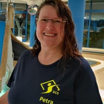 Frau mit nassen Haaren in einem blauen T-Shirt lächelt vor einem Innenbereich mit Wasserrutschen und Wasserattraktionen.