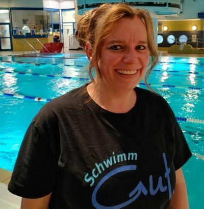 Frau mit fröhlichem Gesicht trägt ein Shirt mit der Aufschrift „SchwimmGut“ vor einem Schwimmbecken.