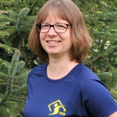 Frau mit blonden, schulterlangen Haaren und Brille, lächelt vor einem Nadelbaum, trägt ein blaues Shirt mit Vereinslogo.