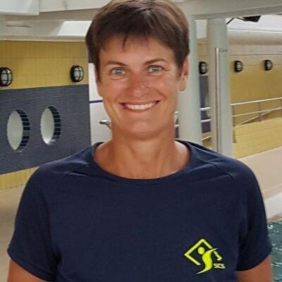 Lächelnde Frau mit kurzer Frisur, trägt ein dunkelblaues Shirt mit gelbem Logo, steht in Schwimmbad-Umgebung.