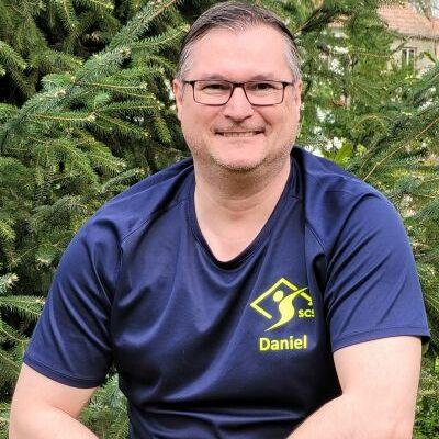 Mann mit Brille und Sportshirt sitzt lächelnd vor einem Hintergrund aus grünen Tannenzweigen.