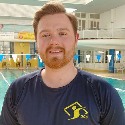 Mann mit rotem Bart und blondem Haar steht in einem Hallenbad vor einem Schwimmbecken mit blauen F&auml;hnchen.
