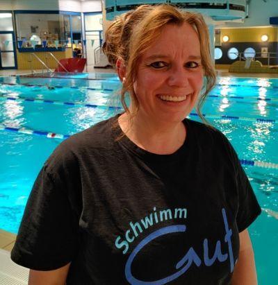 Frau mit fröhlichem Gesicht trägt ein Shirt mit der Aufschrift „SchwimmGut“ vor einem Schwimmbecken.