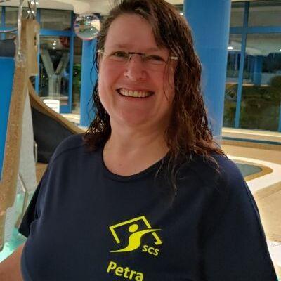 Frau mit nassen Haaren in einem blauen T-Shirt l&auml;chelt vor einem Innenbereich mit Wasserrutschen und Wasserattraktionen.