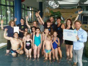 Gruppe von Erwachsenen und Kindern im Schwimmbad h&auml;lt Urkunde, alle jubeln mit erhobenen H&auml;nden.