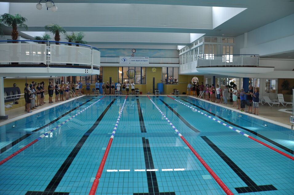 Blick auf ein Schwimmbecken mit bunten Bahnen und Zuschauern an den R&auml;ndern in einem modernen Hallenbad.