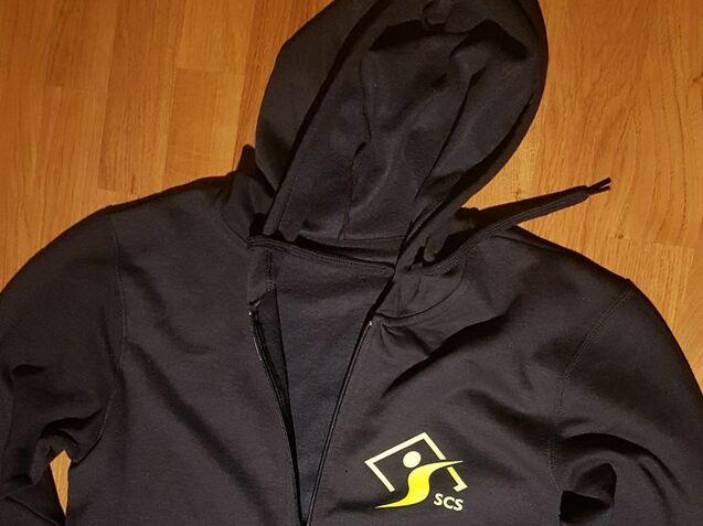 Schwarze Kapuzenjacke mit Reißverschluss und gelbem Logo „SCS“ auf der linken Brusthälfte, auf einem Holztisch ausgelegt.